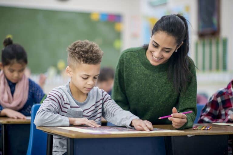 A young boy is indoors in his elementary school classroom. He is sitting at a table and playing a game with the help of his teacher. He is pointing where he wants the teacher to draw.