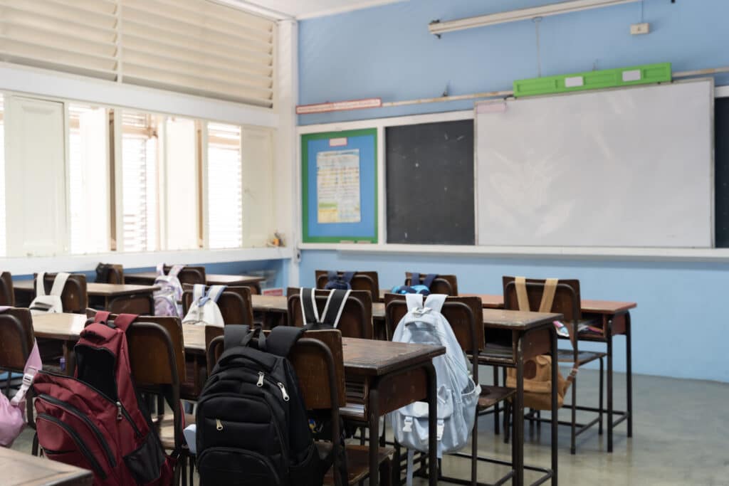 School classroom interior with student backpacks on chairs, wooden desks, blackboard and bright natural light windows