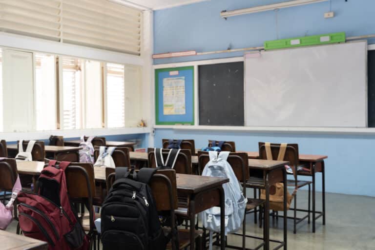 School classroom interior with student backpacks on chairs, wooden desks, blackboard and bright natural light windows